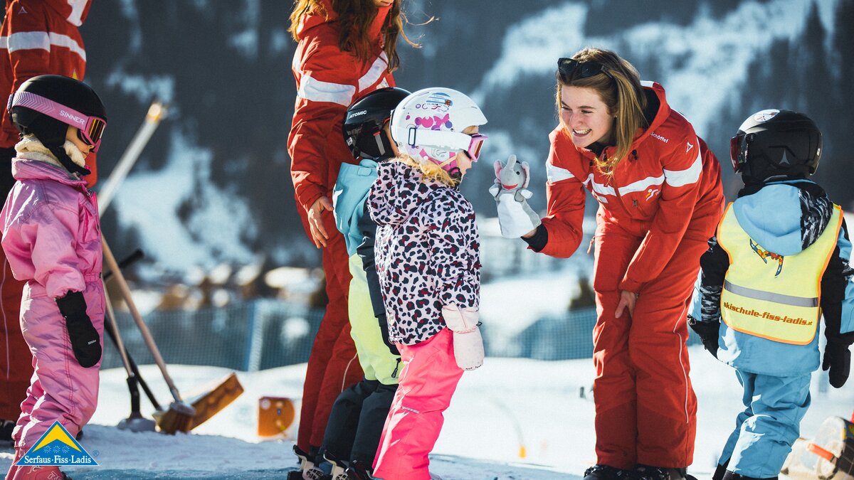Skilehrerin in roter Skischuluniform begrüßt eine Gruppe von Kindern auf Skiern im Schnee; die Kinder tragen bunte Skikleidung und Helme | © Skischule Fiss-Ladis | Maria Knoll