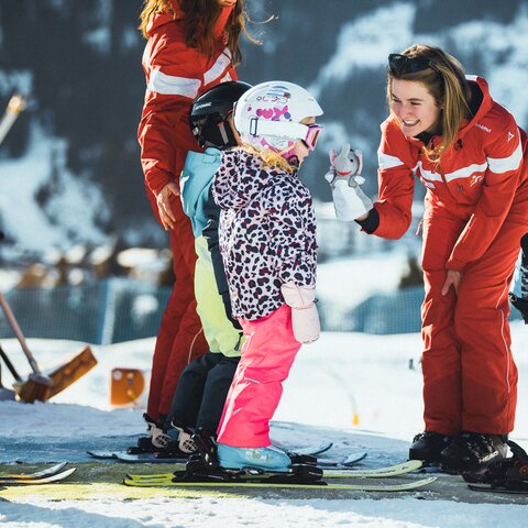Skilehrerin in roter Skischuluniform begrüßt eine Gruppe von Kindern auf Skiern im Schnee; die Kinder tragen bunte Skikleidung und Helme | © Skischule Fiss-Ladis | Maria Knoll