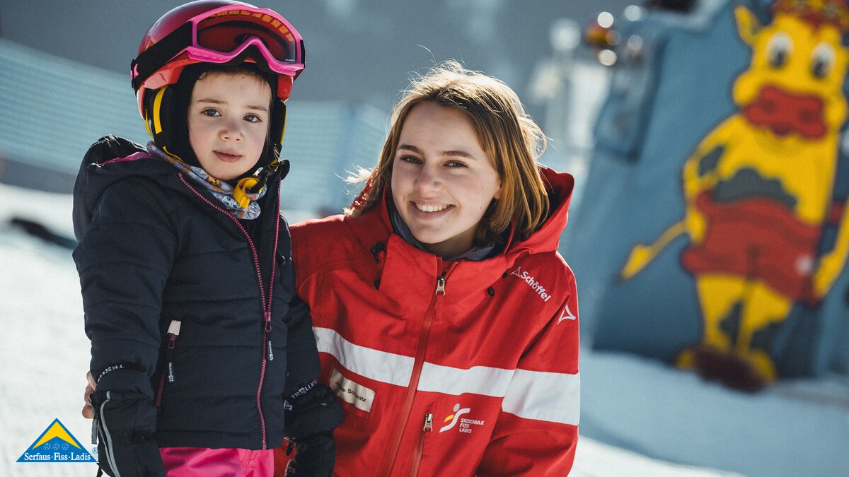 Eine Skilehrerin in roter Skischuluniform steht lächelnd neben einem kleinen Kind mit rosa Helm und Skiausrüstung im Berta's Kinderland. Im Hintergrund ist ein buntes Maskottchen zu sehen. | © Skischule Fiss-Ladis | Maria Knoll