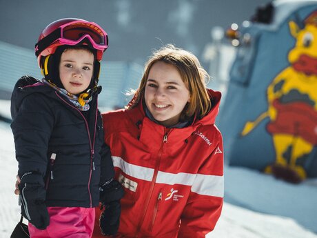 Eine Skilehrerin in roter Skischuluniform steht lächelnd neben einem kleinen Kind mit rosa Helm und Skiausrüstung im Berta's Kinderland. Im Hintergrund ist ein buntes Maskottchen zu sehen. | © Skischule Fiss-Ladis | Maria Knoll