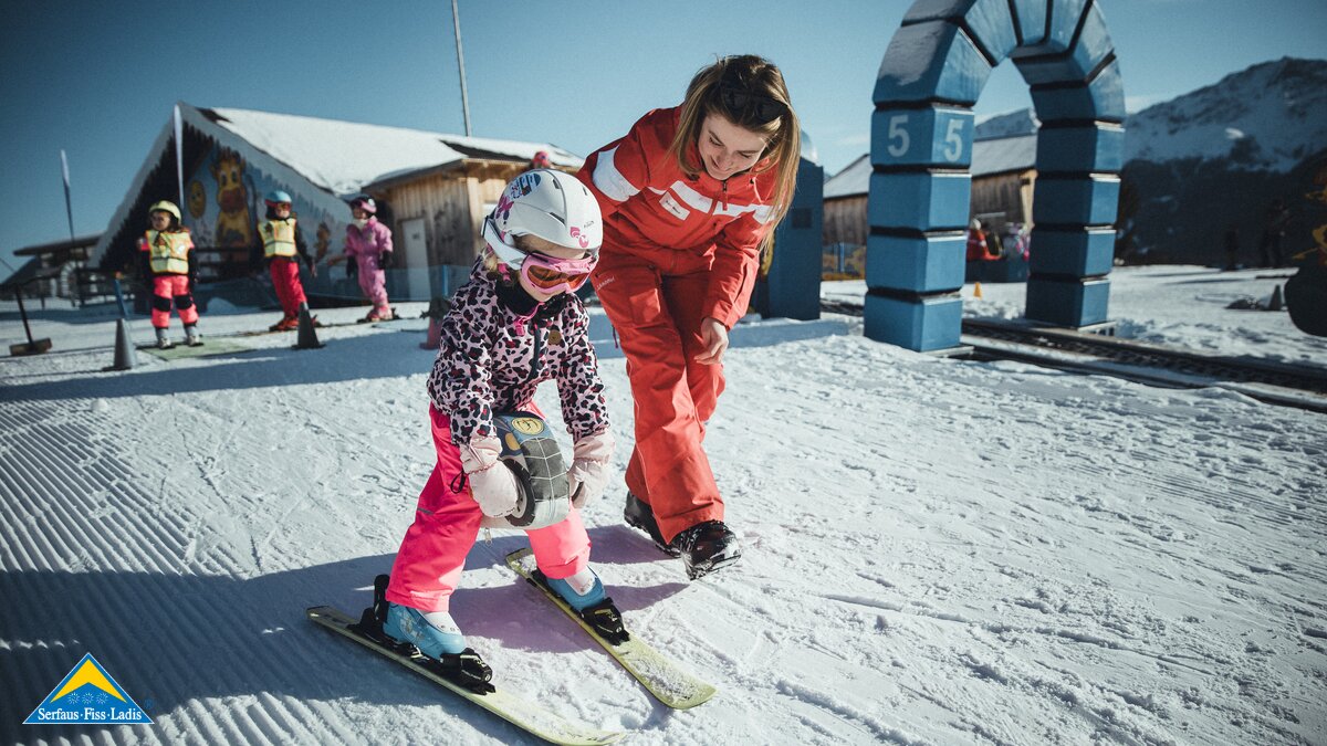 Eine Skilehrerin in roter Skischuluniform unterstützt ein kleines Mädchen beim Skifahren im Berta's Kinderland. Das Mädchen trägt rosa Skihosen, eine gemusterte Jacke und einen weißen Helm. Im Hintergrund sind weitere Kinder in bunter Skiausrüstung zu sehen. | © Skischule Fiss-Ladis | Maria Knoll