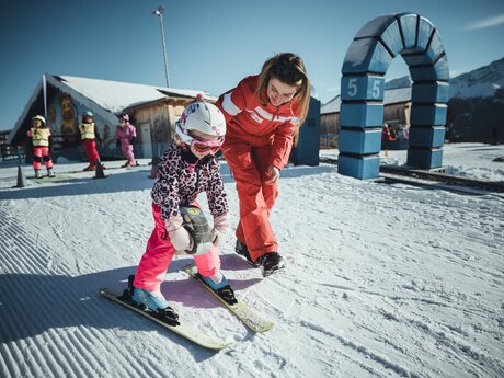 Eine Skilehrerin in roter Skischuluniform unterstützt ein kleines Mädchen beim Skifahren im Berta's Kinderland. Das Mädchen trägt rosa Skihosen, eine gemusterte Jacke und einen weißen Helm. Im Hintergrund sind weitere Kinder in bunter Skiausrüstung zu sehen. | © Skischule Fiss-Ladis | Maria Knoll