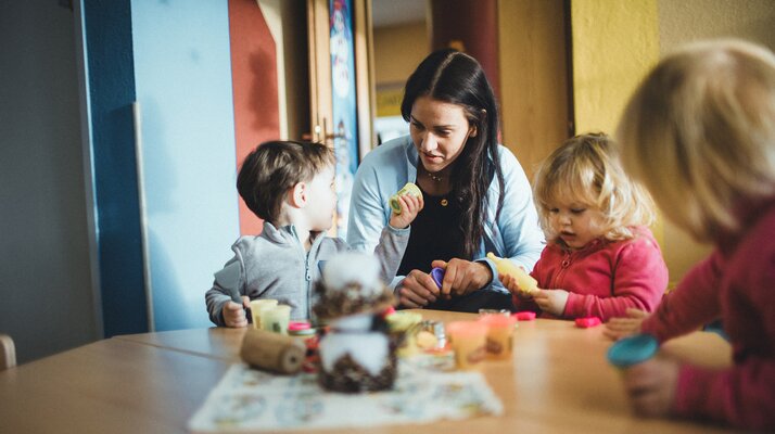 Eine Betreuerin sitzt mit drei kleinen Kindern an einem Tisch in der Murmlikrippe in Serfaus. Die Kinder spielen mit Knete und Bastelmaterial, während die Betreuerin liebevoll mit ihnen interagiert. Im Hintergrund sind bunte Wände und eine Tür zu sehen. | © Skischule Fiss-Ladis | Maria Knoll