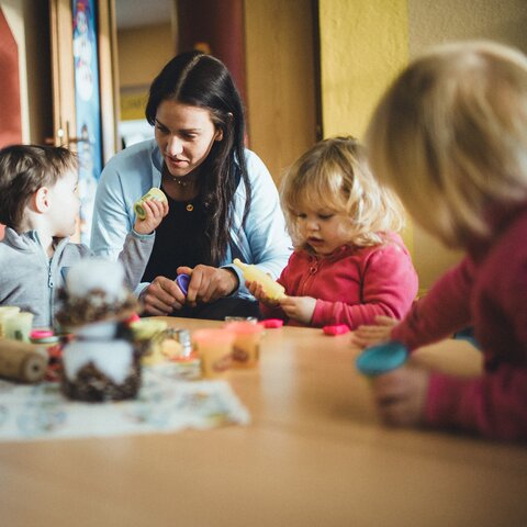 Eine Betreuerin sitzt mit drei kleinen Kindern an einem Tisch in der Murmlikrippe in Serfaus. Die Kinder spielen mit Knete und Bastelmaterial, während die Betreuerin liebevoll mit ihnen interagiert. Im Hintergrund sind bunte Wände und eine Tür zu sehen. | © Skischule Fiss-Ladis | Maria Knoll