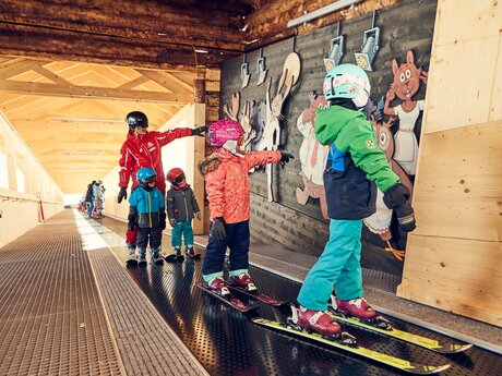 Skilehrerin begleitet Kinder auf einem Förderband beim Skikurs in Serfaus, Wandbilder mit Tieren im Hintergrund | © Skischule Serfaus