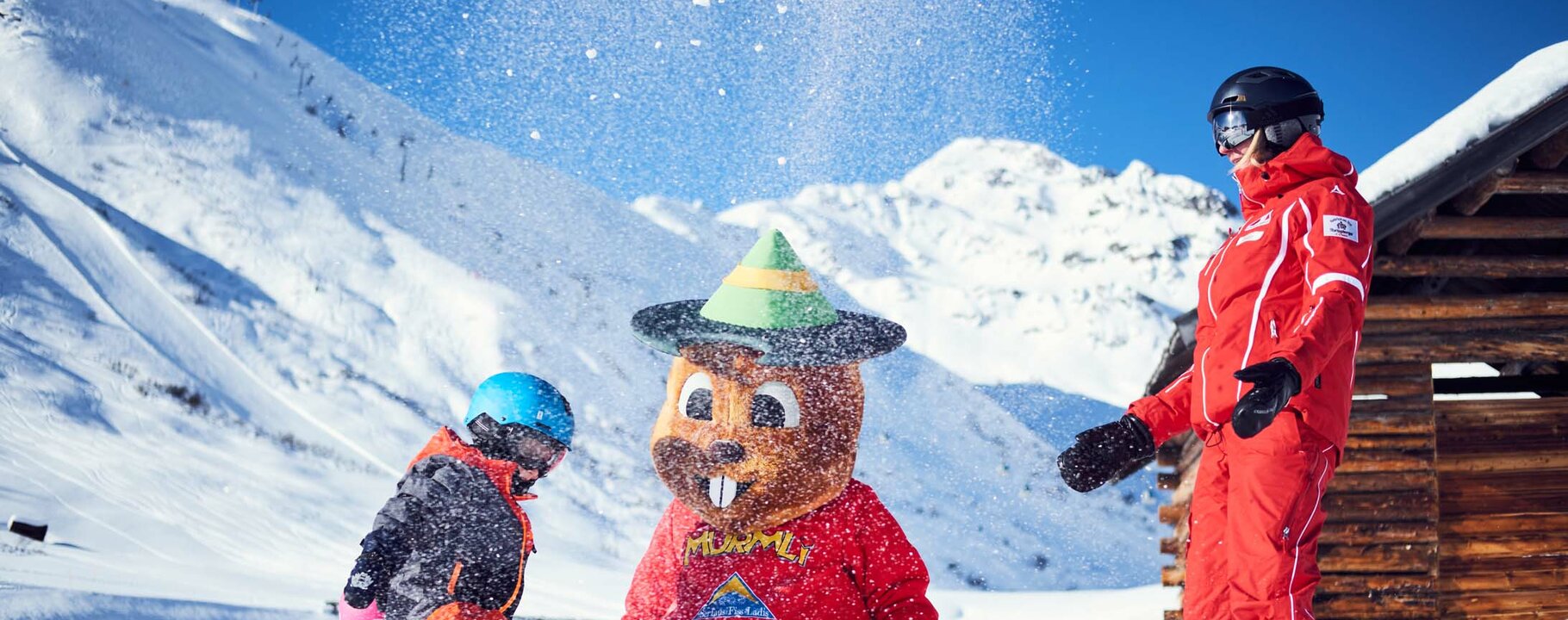 Kind in Skibekleidung und Skilehrerin spielen mit dem Maskottchen Murmli im verschneiten Skigebiet von Serfaus | © christianwaldegger.com