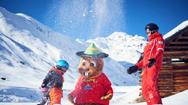 Kind in Skibekleidung und Skilehrerin spielen mit dem Maskottchen Murmli im verschneiten Skigebiet von Serfaus | © christianwaldegger.com