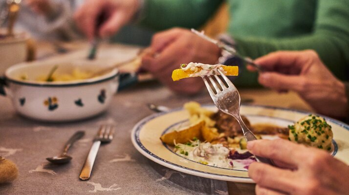 Nahaufnahme eines traditionellen Abendessens in gemütlicher Hüttenatmosphäre mit Knödel, Braten und Beilagen, Hände greifen nach dem Essen | © Seilbahn Komperdell GmbH | Christian Waldegger