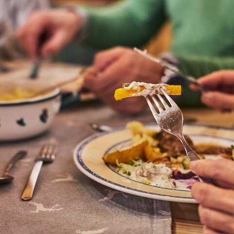 Nahaufnahme eines traditionellen Abendessens in gemütlicher Hüttenatmosphäre mit Knödel, Braten und Beilagen, Hände greifen nach dem Essen | © Seilbahn Komperdell GmbH | Christian Waldegger