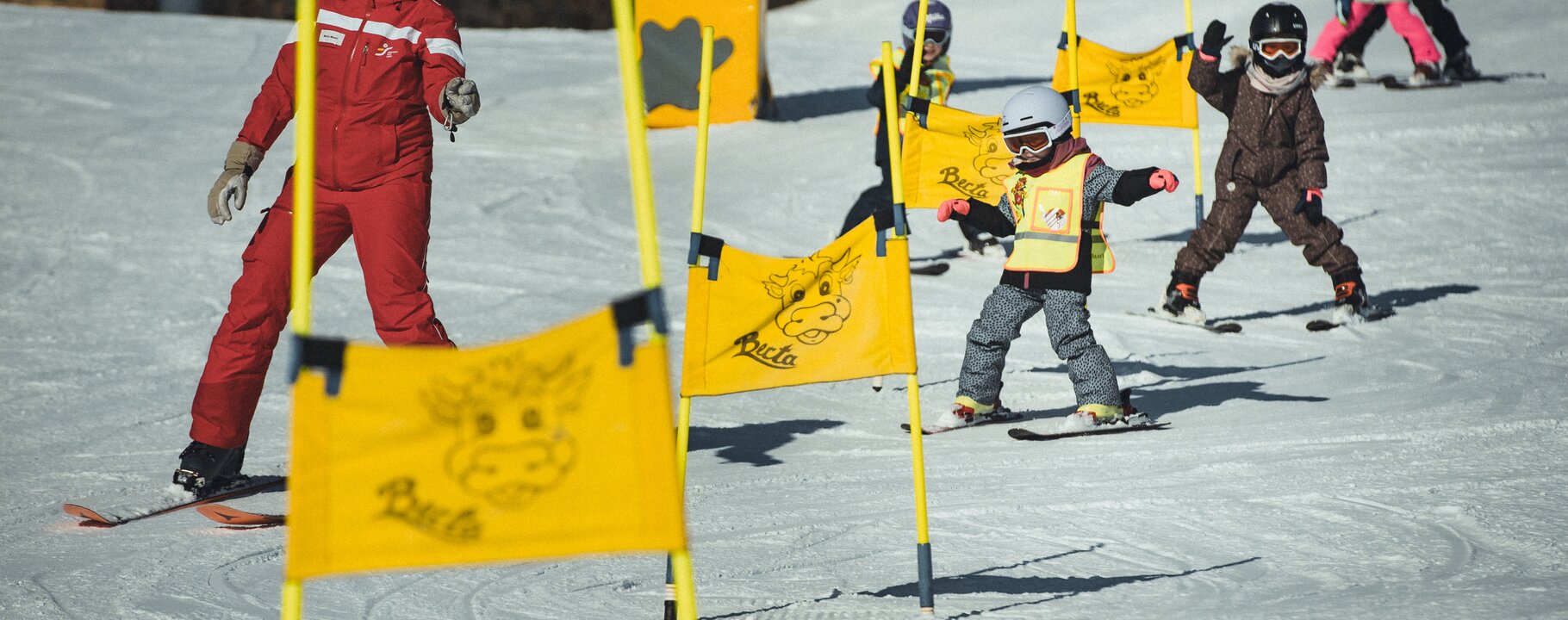 A ski instructor in a red ski school uniform guides a group of children skiing through yellow slalom flags in Berta's Kinderland in Serfaus-Fiss-Ladis | © Skischule Fiss-Ladis | Maria Knoll