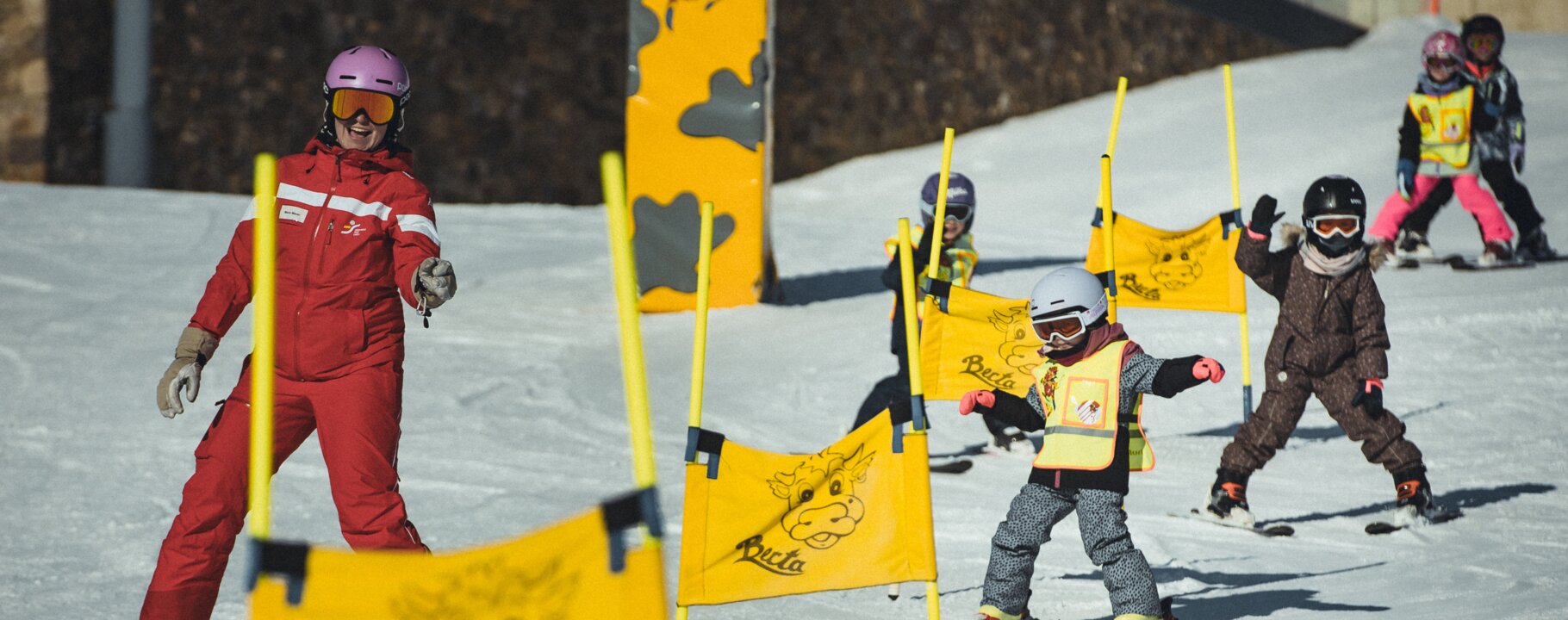 Eine Skilehrerin in roter Skischuluniform leitet eine Gruppe von Kindern beim Skifahren durch gelbe Slalom-Fahnen im Berta's Kinderland in Serfaus-Fiss-Ladis | © Skischule Fiss-Ladis | Maria Knoll