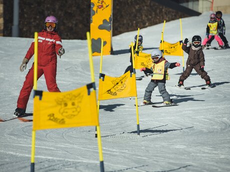 Eine Skilehrerin in roter Skischuluniform leitet eine Gruppe von Kindern beim Skifahren durch gelbe Slalom-Fahnen im Berta's Kinderland in Serfaus-Fiss-Ladis | © Skischule Fiss-Ladis | Maria Knoll