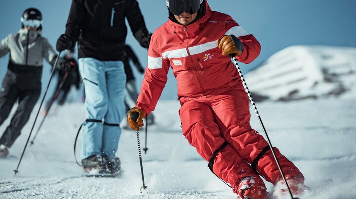 Ein Skilehrer in roter Skischuluniform fährt dynamisch voraus, gefolgt von einer Gruppe von Skifahrern im Schnee; alle tragen Helme und Skibrillen | © Skischule Fiss-Ladis | Maria Knoll