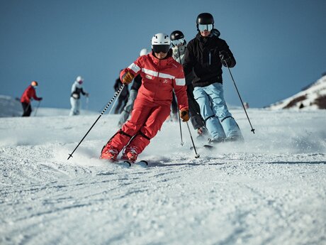 Ein Skilehrer in roter Skiausrüstung führt eine Gruppe von Skifahrern auf einer perfekt präparierten Piste. Die Teilnehmer folgen in sicherem Abstand und üben die Carving-Technik. Im Hintergrund sind weitere Skifahrer zu sehen. | © Skischule Fiss-Ladis | Maria Knoll