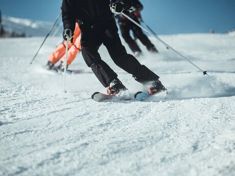 Mehrere Skifahrer in schwarzer und orangefarbener Skiausrüstung fahren in schneller Carving-Technik über eine verschneite Piste. Der Fokus liegt auf den Skiern und den dynamischen Bewegungen im Schnee. | © Skischule Fiss-Ladis | Maria Knoll