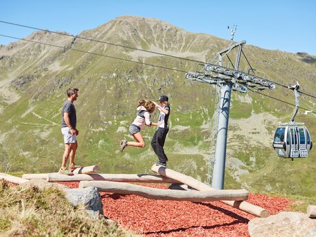 Zwei Jugendliche und ein Erwachsener balancieren auf Holzstämmen unterhalb einer Seilbahn vor beeindruckender Bergkulisse in Serfaus-Fiss-Ladis | © Christian Waldegger
