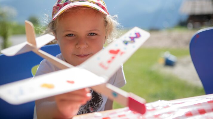 Kleines Mädchen hält stolz ein selbstbemaltes Modellflugzeug beim Basteln im Freien in Serfaus-Fiss-Ladis | © Serfaus-Fiss-Marketing GmbH | Andreas Kirschner