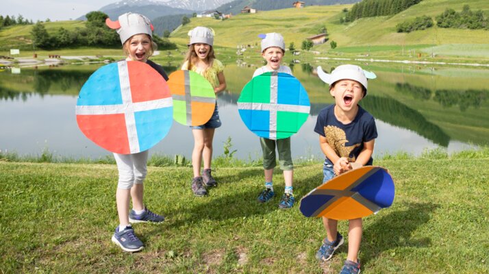 Vier lachende Kinder mit Wikingerhelmen und bunten Schilden beim Högsee in Serfaus-Fiss-Ladis, umgeben von grüner Natur und Bergen | © Andreas Kirschner