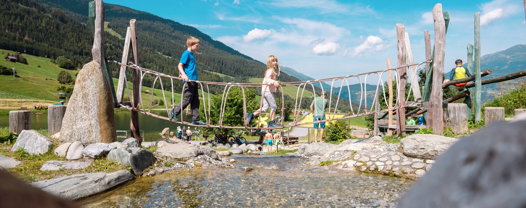 Zwei Kinder balancieren über eine Seilbrücke über einen Bach im Hög Adventure Park in Serfaus-Fiss-Ladis, umgeben von grünen Wiesen und Bergen | © Serfaus-Fiss-Marketing GmbH | Fabian Schrigi