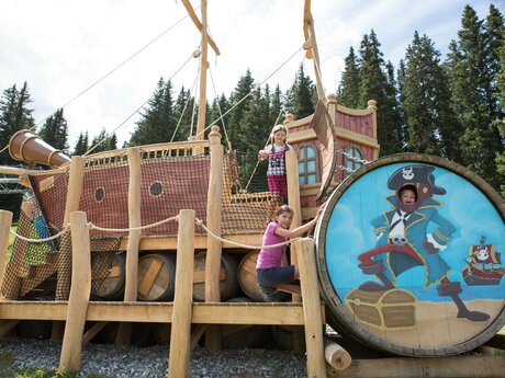 Zwei Kinder klettern auf ein großes hölzernes Piratenschiff am Piratenweg in Serfaus-Fiss-Ladis, ein weiteres Kind schaut lachend durch ein Fotofenster mit Piratenmotiv | © Serfaus-Fiss-Ladis Marketing GmbH | Andreas Kirschner