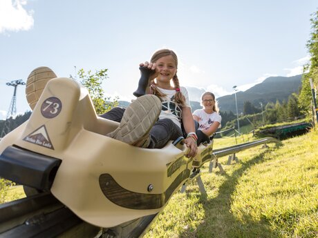 Zwei Mädchen fahren lachend mit dem Fisser Flitzer den Rodelbahnhang hinunter, im Hintergrund grüne Wiesen und Berge | © Serfaus-Fiss-Ladis Marketing GmbH