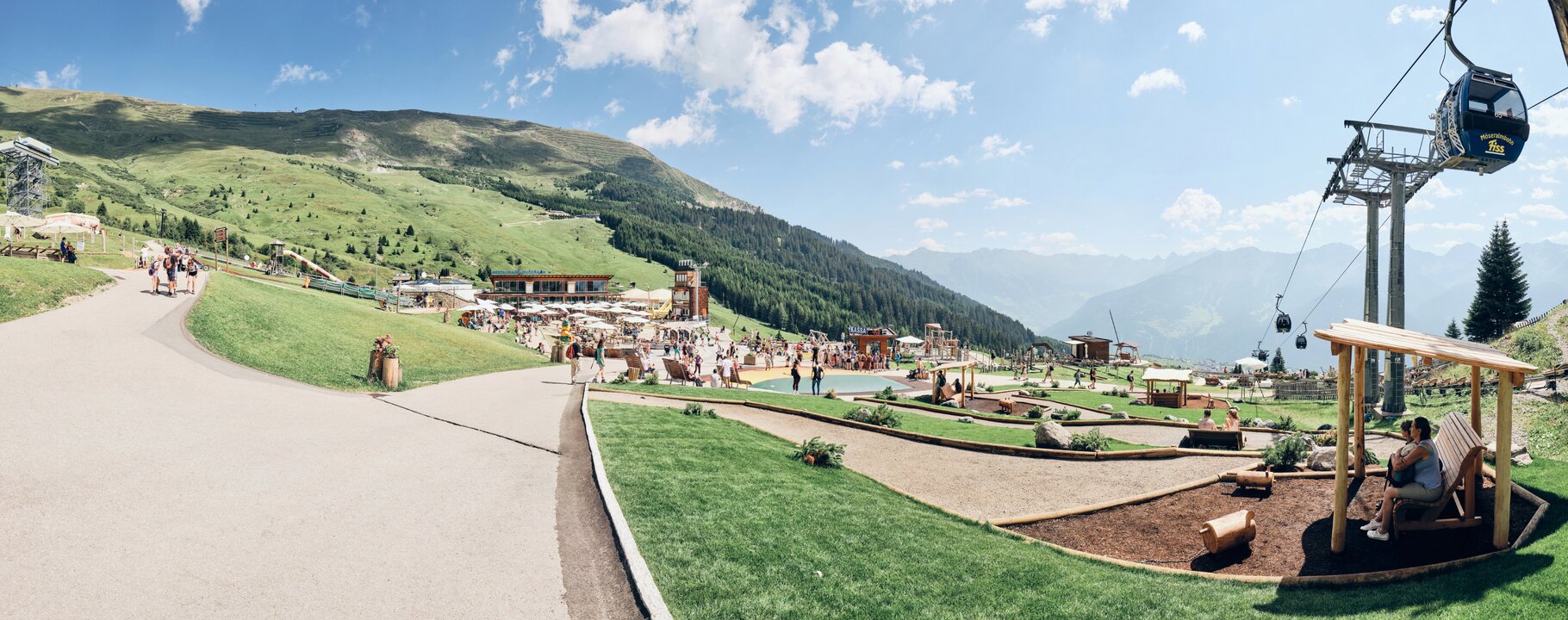 Panoramaaufnahme vom Sommer-Funpark Fiss in Serfaus-Fiss-Ladis mit spielenden Kindern, der Möseralmbahn und entspannten Besuchern unter einem Holzunterstand | © Fisser Bergbahnen GmbH