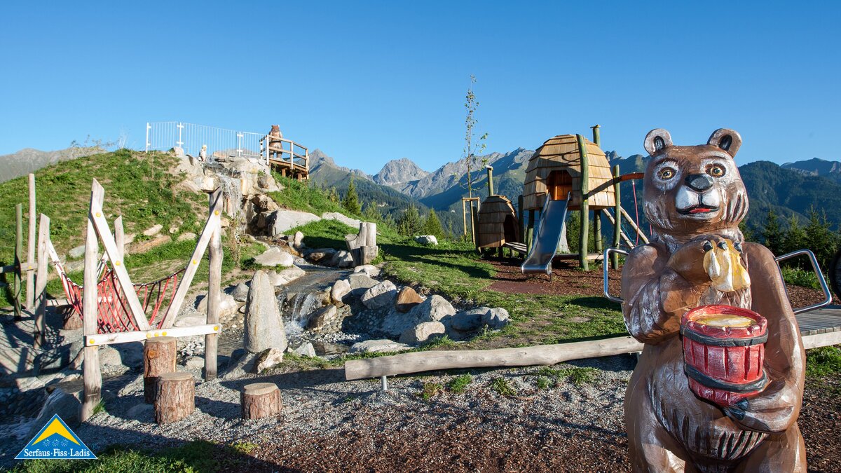 Spielplatz mit Kletterbereich, Rutsche, Hängebrücke und Holzfigur eines Bären mit Honigtopf vor Bergkulisse in Serfaus-Fiss-Ladis | © Serfaus-Fiss-Ladis Marketing GmbH