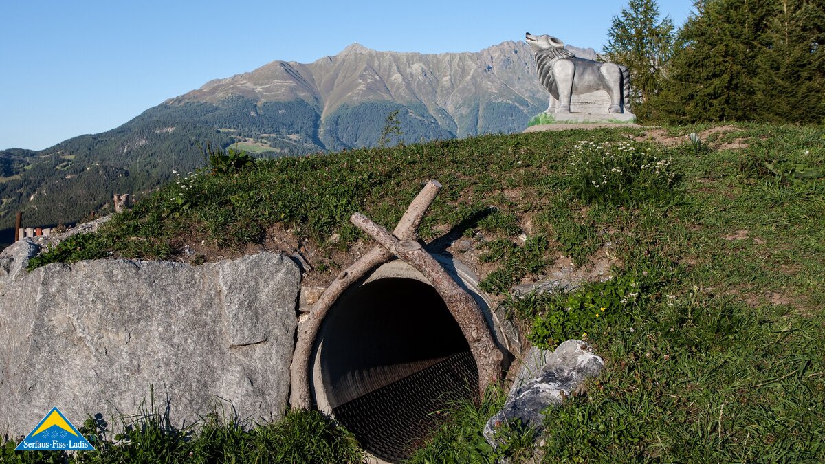 Röhrentunnel im Gras mit Holzeingang und einer großen Wolfsstatue auf dem Hügel im Hintergrund, inmitten alpiner Landschaft | © Serfaus-Fiss-Ladis Marketing GmbH