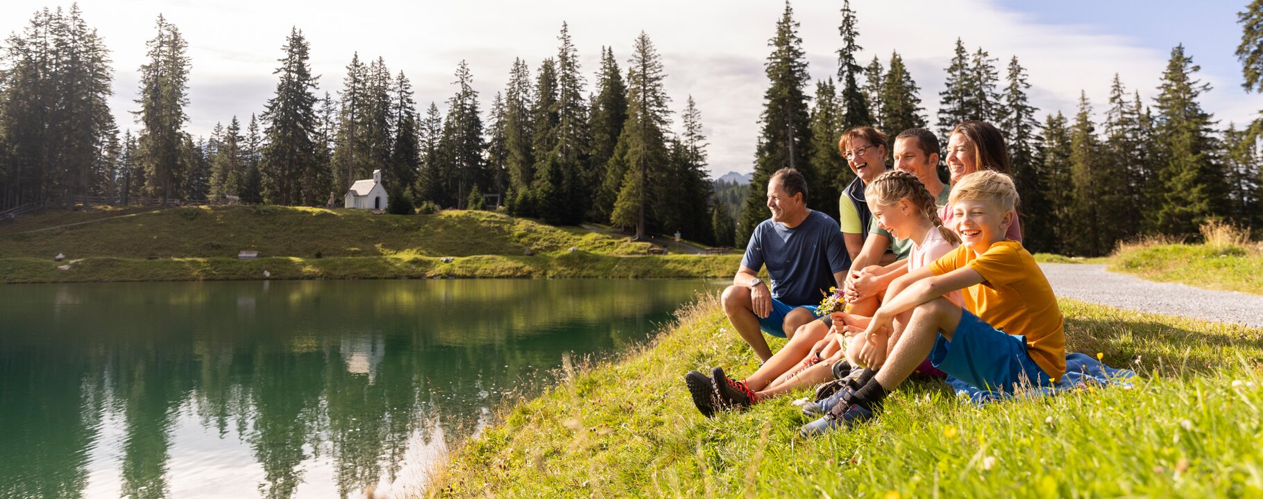 Mehrere Familienmitglieder sitzen lachend am Ufer des Högsees in Serfaus-Fiss-Ladis, mit Blick auf das spiegelnde Wasser und umliegende Wälder | © Serfaus-Fiss-Ladis Marketing GmbH | Daniel Zangerl