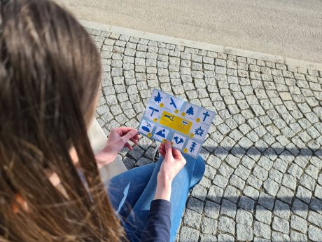 Mädchen hält eine Stempelkarte in der Hand und sitzt auf einem gepflasterten Weg beim Dorfrundgang | © Serfaus-Fiss-Ladis Marketing GmbH