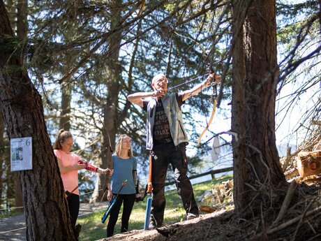 Drei Personen beim Bogenschießen auf einem Waldparcours in Serfaus-Fiss-Ladis, ein Trainer zeigt die richtige Schusstechnik | © Serfaus-Fiss-Ladis Marketing GmbH | Andreas Kirschner