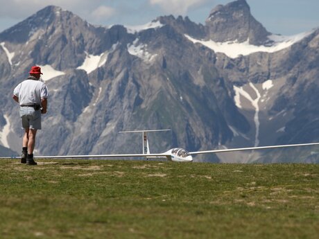Mann mit rotem Hut geht auf einen Modellsegelflieger in Serfaus-Fiss-Ladis zu, im Hintergrund hohe Berge mit Schneefeldern | © Serfaus-Fiss-Ladis Marketing GmbH
