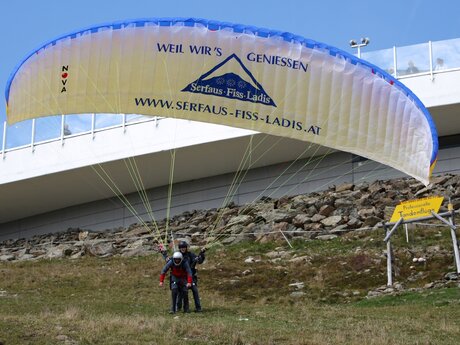 Zwei Personen beim Start zum Tandem-Paragleiten in Serfaus-Fiss-Ladis, Paraglider mit Werbeaufschrift im Hintergrund moderne Station | © Serfaus-Fiss-Ladis Marketing GmbH