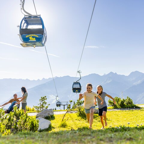 Zwei Kinder rennen barfuß über eine Wiese im Sommer Funpark Fiss, darüber schwebt die Möseralmbahn, im Hintergrund entspannen Erwachsene in Liegestühlen und Hängematten vor alpinem Panorama | © Serfaus-Fiss-Ladis Marketing GmbH | Andreas Kirschner