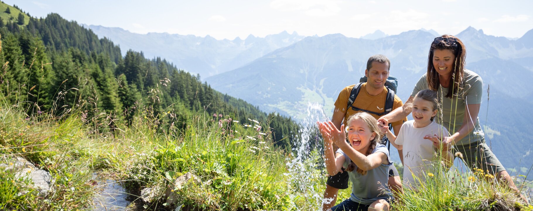 Kinder spielen lachend im klaren Wasser eines Gebirgsbachs, während die Eltern zusehen – Familienerlebnis in der Natur von Serfaus-Fiss-Ladis mit Panoramablick auf die Alpen | © Serfaus-Fiss-Ladis Marketing GmbH | Andreas Kirschner