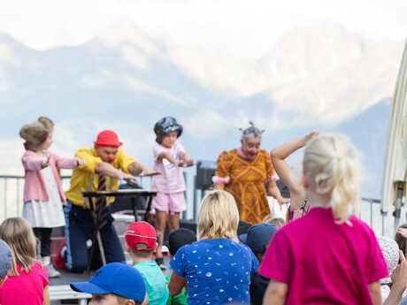 Children watching a fun show with clowns and dressed-up kids on a stage with a mountain backdrop | © Serfaus-Fiss-Ladis Marketing GmbH | Andreas Kirschner