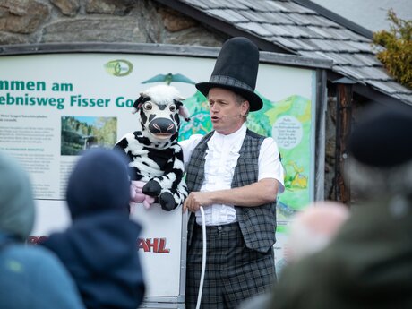 A ventriloquist in traditional clothing entertains an audience with a cow puppet at the Fisser Gonde adventure trail | © Serfaus-Fiss-Ladis Marketing GmbH | Andreas Kirschner