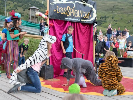 Children in animal costumes perform in front of a red curtain at an event called "Das Spielvolk" with spectators watching | © Serfaus-Fiss-Ladis Marketing GmbH