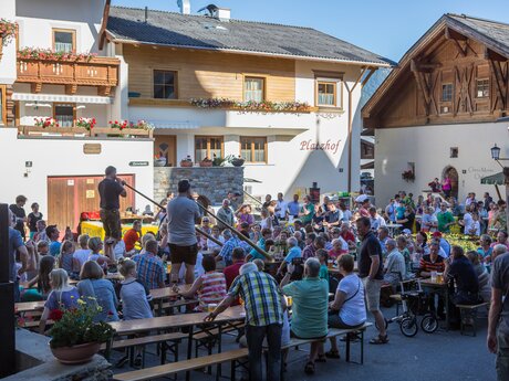Zahlreiche Besucher feiern das Fest am Brunnen auf einem belebten Dorfplatz, während Musiker traditionelle Alphörner spielen | © Serfaus-Fiss-Ladis Marketing GmbH | Andreas Kirschner