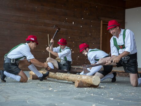Vier Männer in Tracht und roten Hüten hacken bei einer Vorführung gemeinsam Holzstämme | © Serfaus-Fiss-Ladis Marketing GmbH | Andreas Kirschner