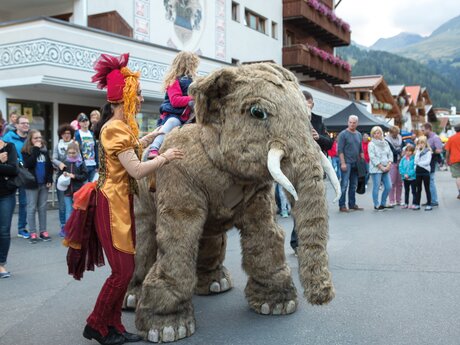 Ein Kind reitet auf einem Mammut-Kostüm, begleitet von einer Künstlerin, während Zuschauer bei einem Straßenfest zusehen | © Serfaus-Fiss-Ladis Marketing GmbH | Andreas Kirschner
