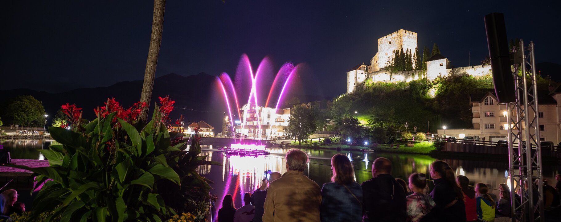 Zuschauer sitzen am Ufer und beobachten eine bunte Wassershow vor einer beleuchteten Burg bei Nacht | © Serfaus-Fiss-Ladis Marketing GmbH | Andreas Kirschner