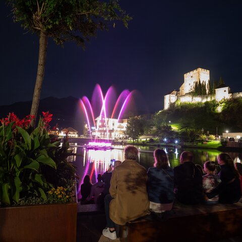 Zuschauer sitzen am Ufer und beobachten eine bunte Wassershow vor einer beleuchteten Burg bei Nacht | © Serfaus-Fiss-Ladis Marketing GmbH | Andreas Kirschner