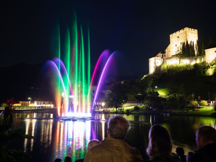 Beleuchtete Wassershow mit lila Fontänen beim Event „Magic Ladis“ am Lader Weiher, im Hintergrund die beleuchtete Burg Laudeck bei Nacht | © Serfaus-Fiss-Ladis Marketing GmbH | Andreas Kirschner