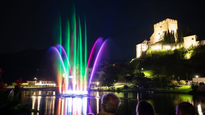 Beleuchtete Wassershow mit lila Fontänen beim Event „Magic Ladis“ am Lader Weiher, im Hintergrund die beleuchtete Burg Laudeck bei Nacht | © Serfaus-Fiss-Ladis Marketing GmbH | Andreas Kirschner