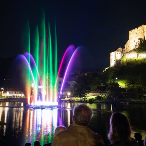 Beleuchtete Wassershow mit lila Fontänen beim Event „Magic Ladis“ am Lader Weiher, im Hintergrund die beleuchtete Burg Laudeck bei Nacht | © Serfaus-Fiss-Ladis Marketing GmbH | Andreas Kirschner