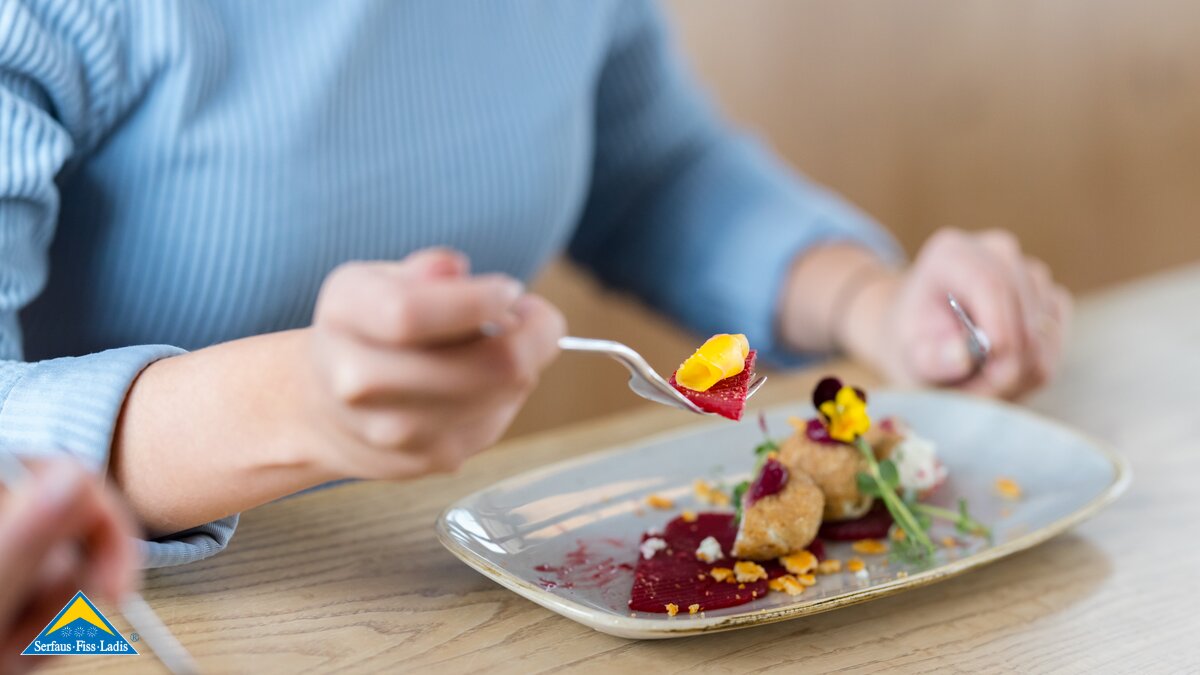 Frau in hellblauem Pullover isst ein kunstvoll angerichtetes Gericht mit Roter Bete und essbaren Blüten | © Serfaus-Fiss-Ladis Marketing GmbH | Fabian Schirgi