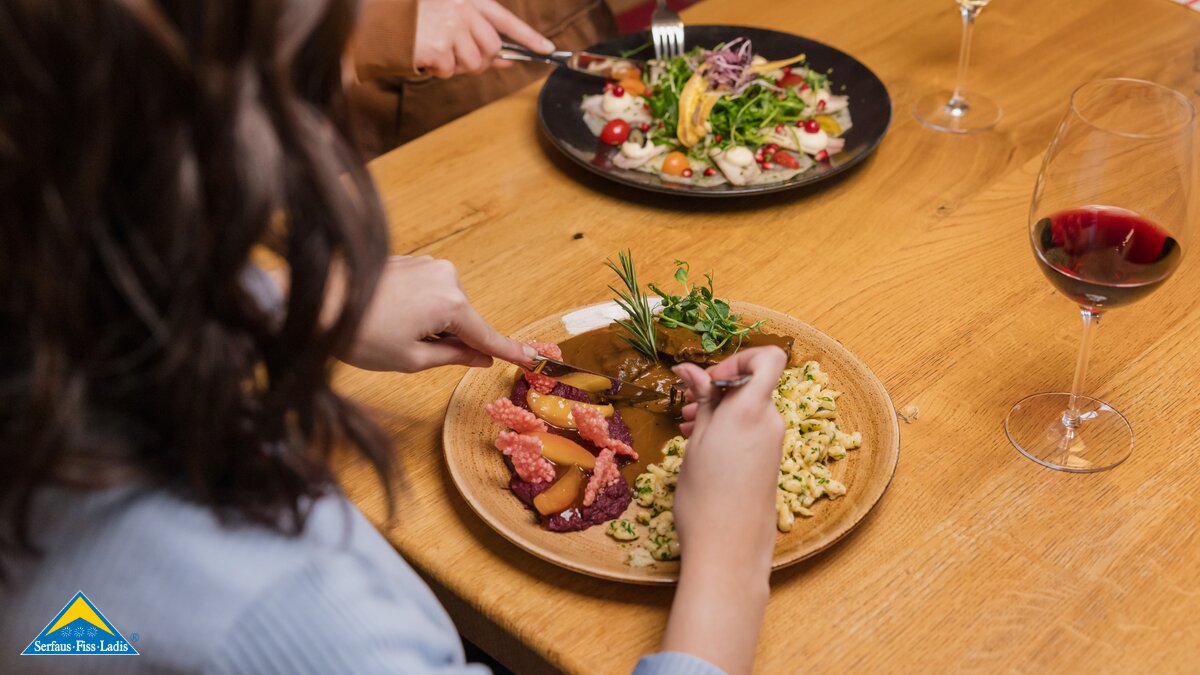 Zwei Personen essen ein Abendessen mit Salat, Fleischgericht, Spätzle und Rotwein an einem Holztisch | © Serfaus-Fiss-Ladis Marketing GmbH | Fabian Schirgi