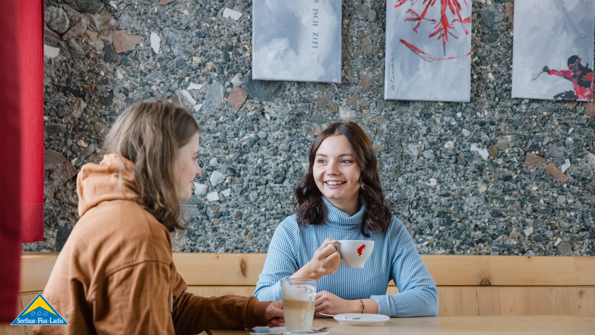 Zwei junge Erwachsene sitzen an einem Holztisch in einem Café, unterhalten sich und trinken Kaffee, im Hintergrund moderne Wandkunst | © Serfaus-Fiss-Ladis Marketing GmbH | Fabian Schirgi