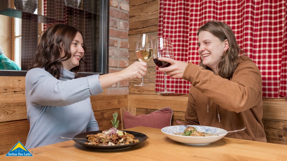 Zwei junge Frauen sitzen in einer gemütlichen Stube, stoßen mit Rot- und Weißwein an und genießen ihr Essen | © Serfaus-Fiss-Ladis Marketing GmbH | Fabian Schirgi